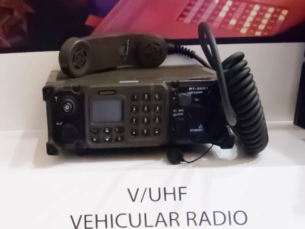 Σκούρος πράσινος ασύρματος οχημάτων V/UHF με πληκτρολόγιο, μικρή οθόνη και ακουστικό με σπιράλ καλώδιο ακουμπισμένο επάνω του, σε λευκό ράφι έκθεσης