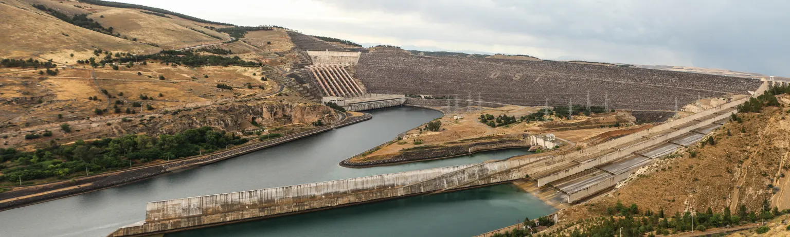 Un long barrage en terre à travers une large vallée, avec un évacuateur de crues en béton au centre et le fleuve qui s’écoule en aval