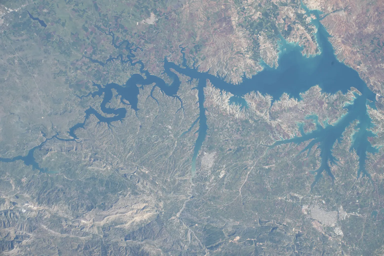 Une vue depuis l’espace d’une grande retenue aux nombreux bras allongés entre des collines brunes, avec le barrage à une extrémité