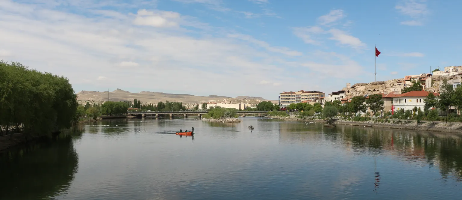 Le Kızılırmak à Avanos : une vaste étendue d’eau calme avec une petite barque orange, un pont routier au loin, des saules sur la rive gauche et des maisons sur la droite sous un ciel bleu