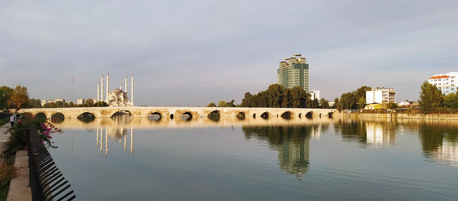 Un large fleuve calme avec un vieux pont de pierre à nombreuses arches, une mosquée et une tour d’habitation sur la rive