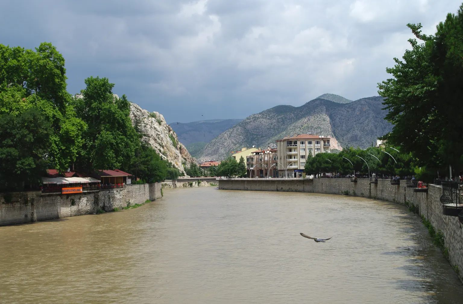 Un fleuve entre des quais de pierre dans une ville, avec des maisons sur la rive et des collines verdoyantes à l’arrière-plan
