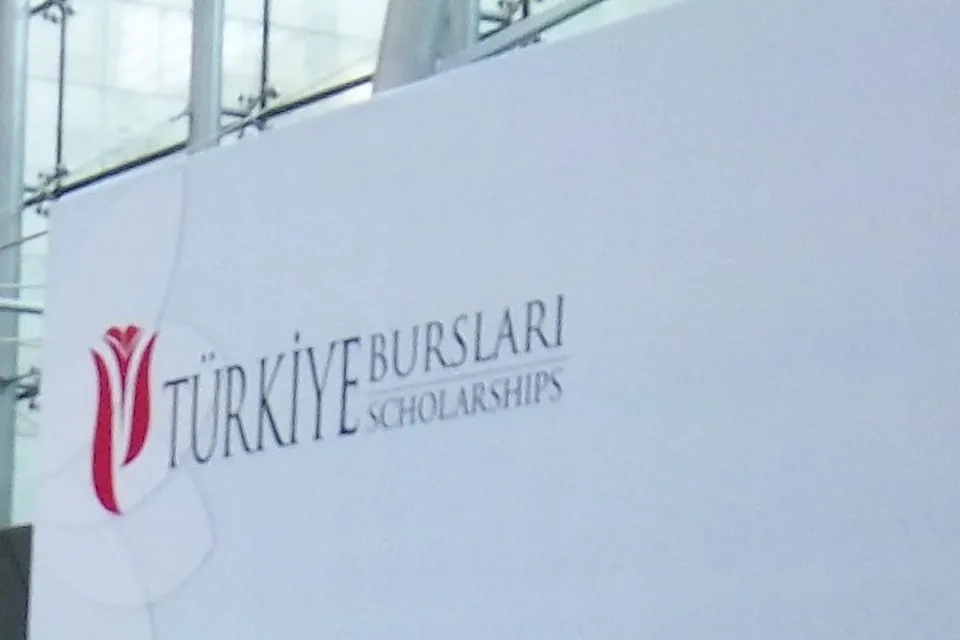 Uma grande parede branca em um aeroporto, com o logotipo de uma tulipa vermelha e as palavras Türkiye Bursları Scholarships em letras escuras, com painéis de vidro acima