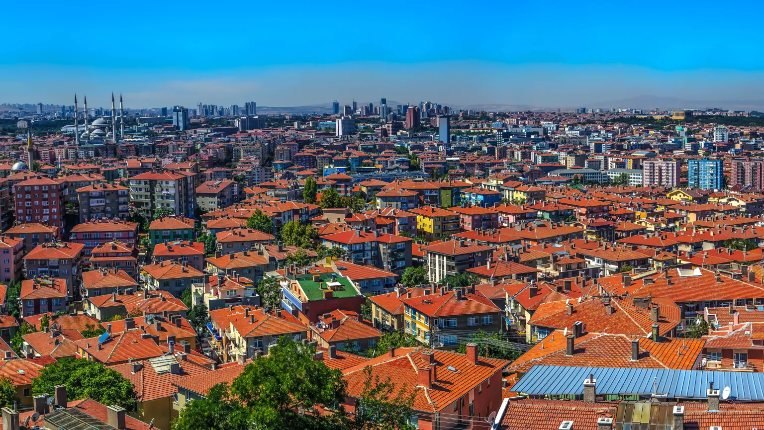 Vista ampla dos telhados de telhas vermelhas e dos blocos de apartamentos de Ancara sob um céu azul, com os minaretes de uma grande mesquita no horizonte à esquerda