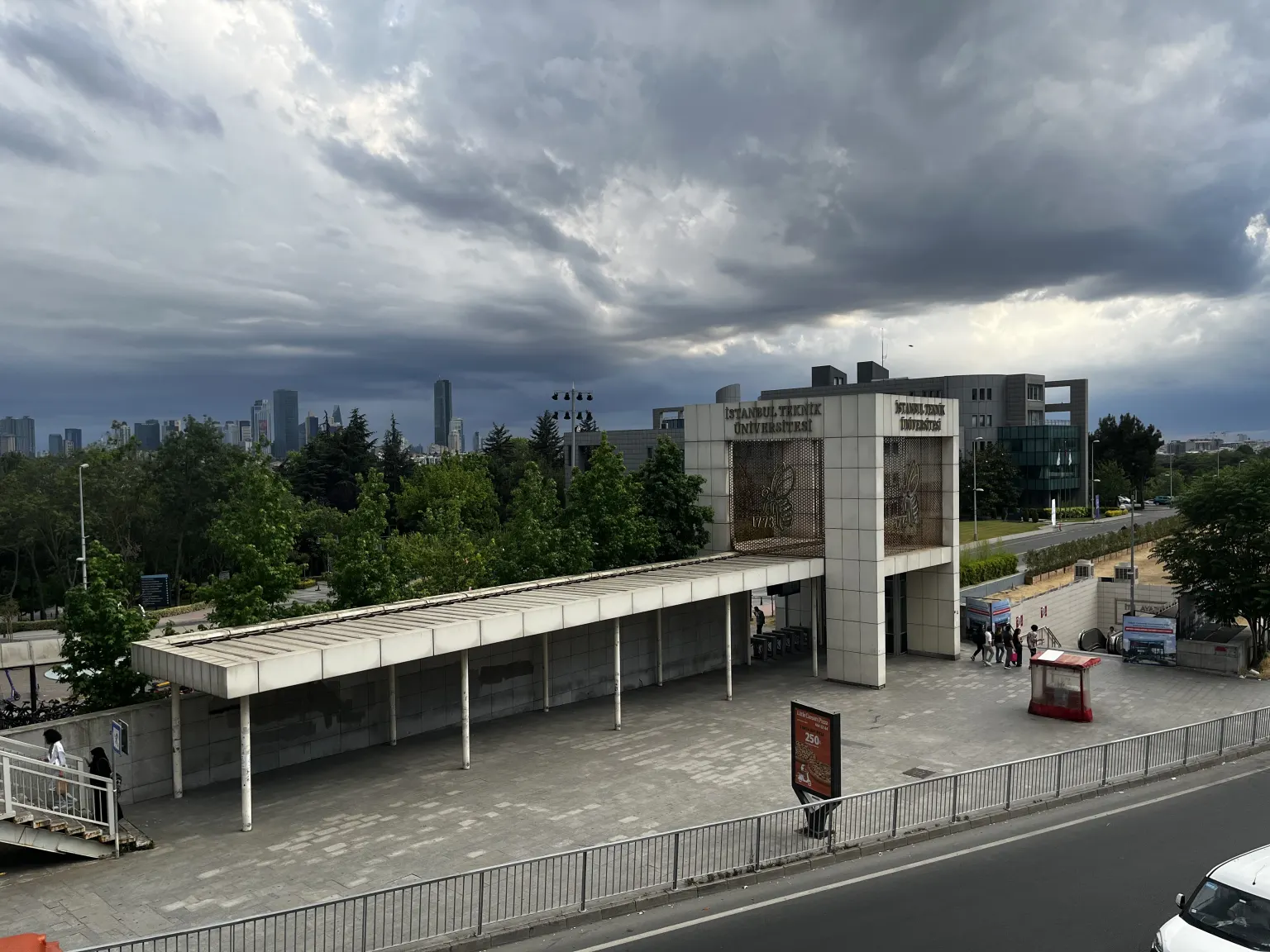 O prédio de entrada da Universidade Técnica de Istambul, um portão de concreto com o nome da universidade e uma longa marquise, sob um céu escuro e nublado, com torres da cidade ao fundo