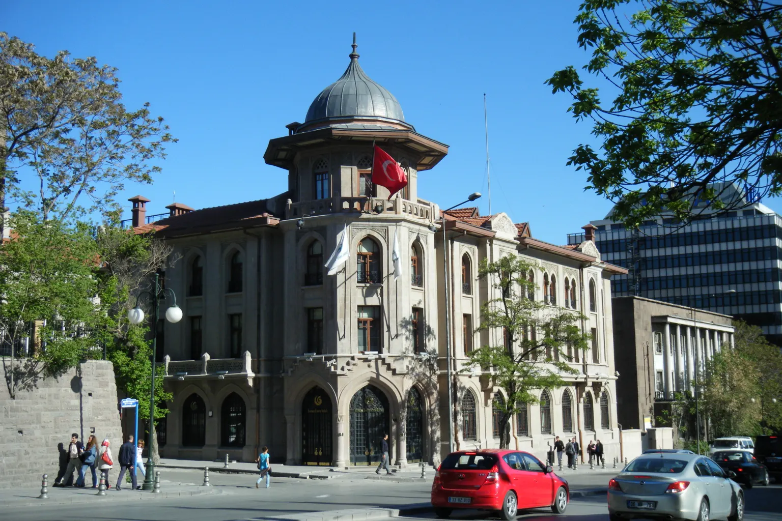 Um prédio de pedra com uma torre de esquina com cúpula e uma bandeira turca em uma esquina de Ancara, com árvores e carros estacionados à frente